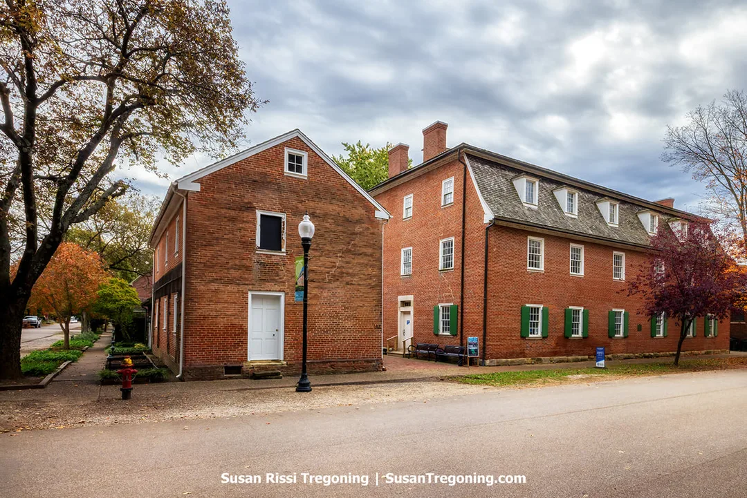 Community House No. 2 in New Harmony, Indiana, stands as a large three‑story brick structure with green shutters and dormer windows, set along a quiet tree‑lined street. A smaller brick building sits beside it, and a vintage-style streetlamp, benches, and autumn foliage contribute to the historic, village‑like atmosphere.