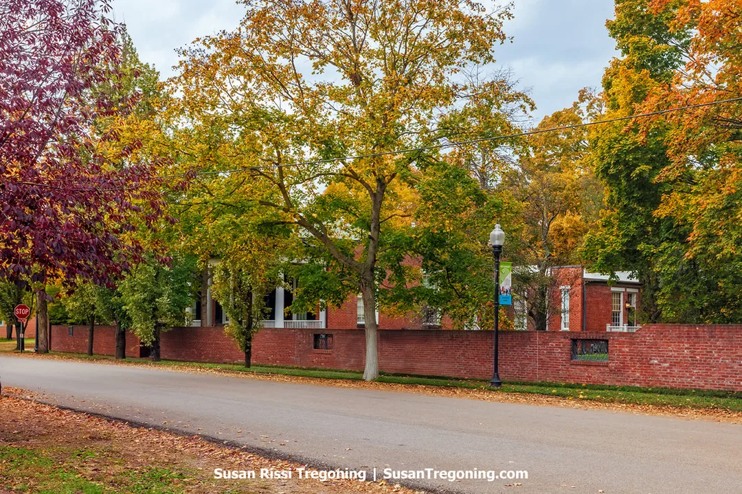 A quiet tree‑lined street in autumn leads past a long red‑brick wall, with the Rapp‑Maclure‑Owen Home partially visible behind the trees. The historic brick house shows white columns and tall windows framed by vibrant yellow, orange, and red foliage. A black streetlamp with a colorful banner stands near the wall, and a stop sign is visible in the distance, giving the scene a calm neighborhood feel.