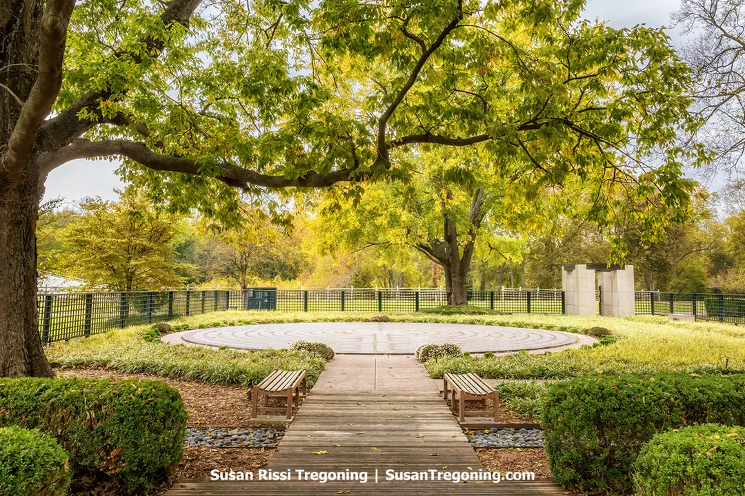 A circular stone labyrinth sits at the center of the Sacred Garden in New Harmony, Indiana, surrounded by autumn trees with golden and rust‑colored leaves. A wooden walkway leads to the labyrinth and is flanked by two benches, while a black metal fence and a simple stone structure frame the quiet, contemplative space.