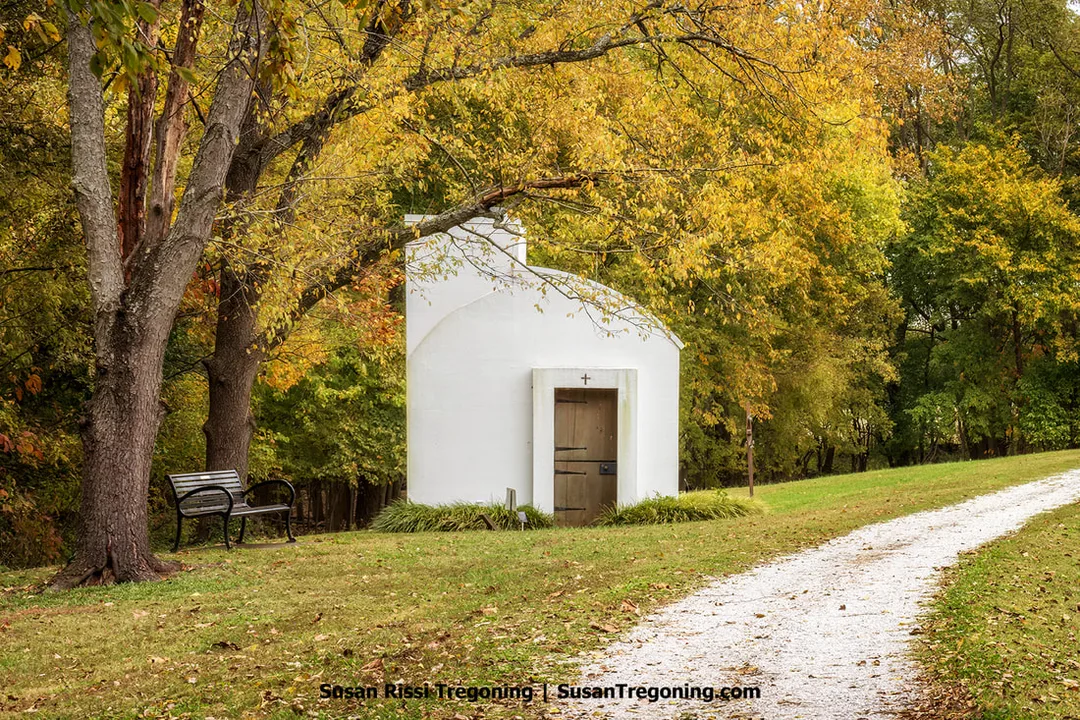 A small white chapel known as the Chapel of the Little Portion stands in a wooded clearing, its simple arched wooden door topped by a cross. A curved gravel path leads to the entrance, and a black metal bench sits beneath an autumn‑colored tree to the left. Tall trees with yellow and orange foliage surround the chapel, creating a quiet, contemplative fall setting.