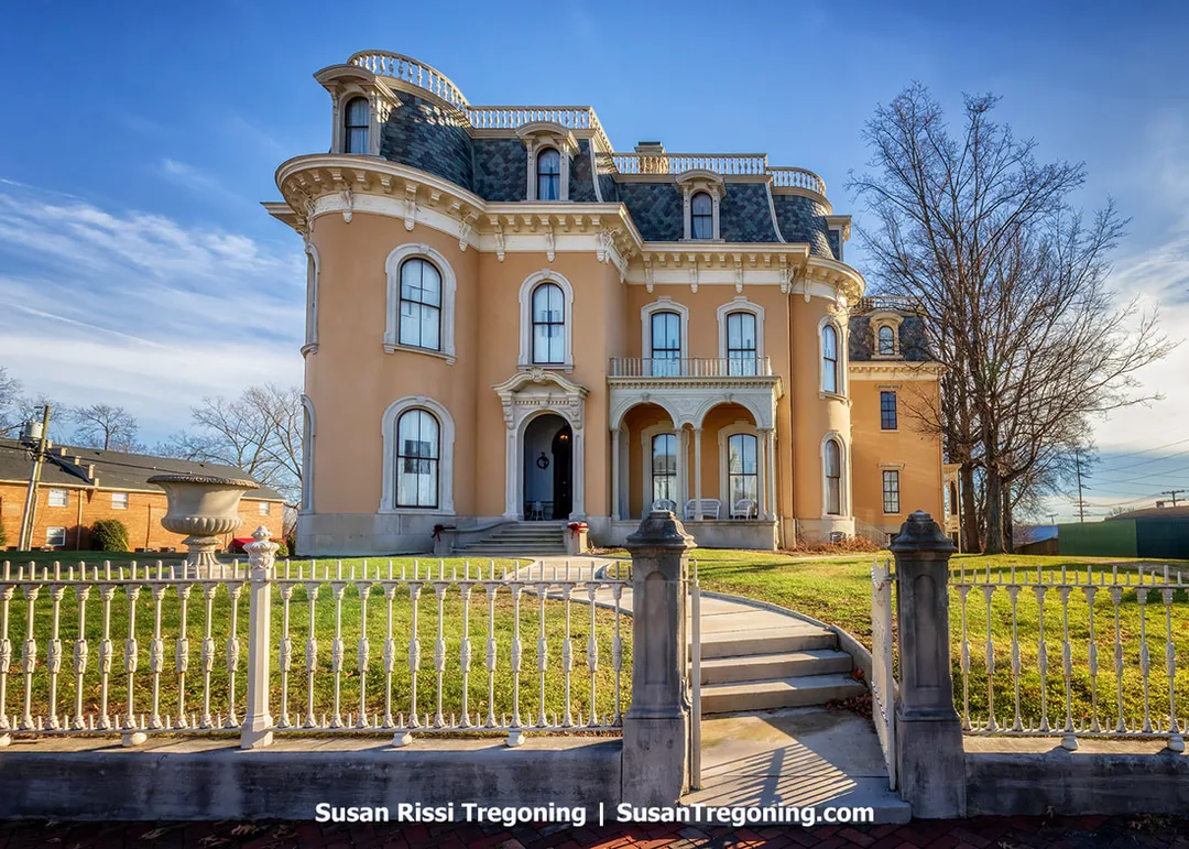 A large Victorian mansion with ornate architectural detailing rises behind a wrought‑iron fence. The multi‑story brick home features tall windows, decorative trim, and a prominent tower, with bare trees framing the historic structure.