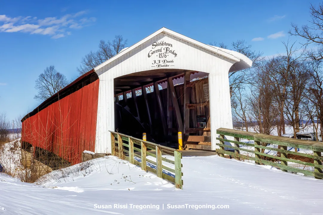 A scenic winter view of the historic Shieldstown Covered Bridge in Jackson County, Indiana. Built in 1876 by J.J. Daniels, this red and white wooden bridge is surrounded by snow under a bright blue sky.