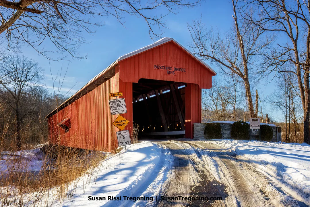 A red wooden covered bridge labeled ‘Busching Bridge 1885’ sits in a snowy, wooded landscape. A snow‑covered road leads to the bridge, and several warning and height‑limit signs stand at the entrance.