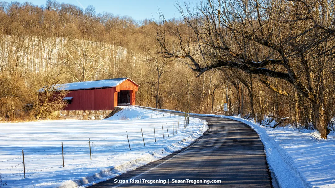 Busching Covered Bridge viewed from a distance across a snow‑covered field, with the road curving upward toward the wooden span in Versailles State Park.