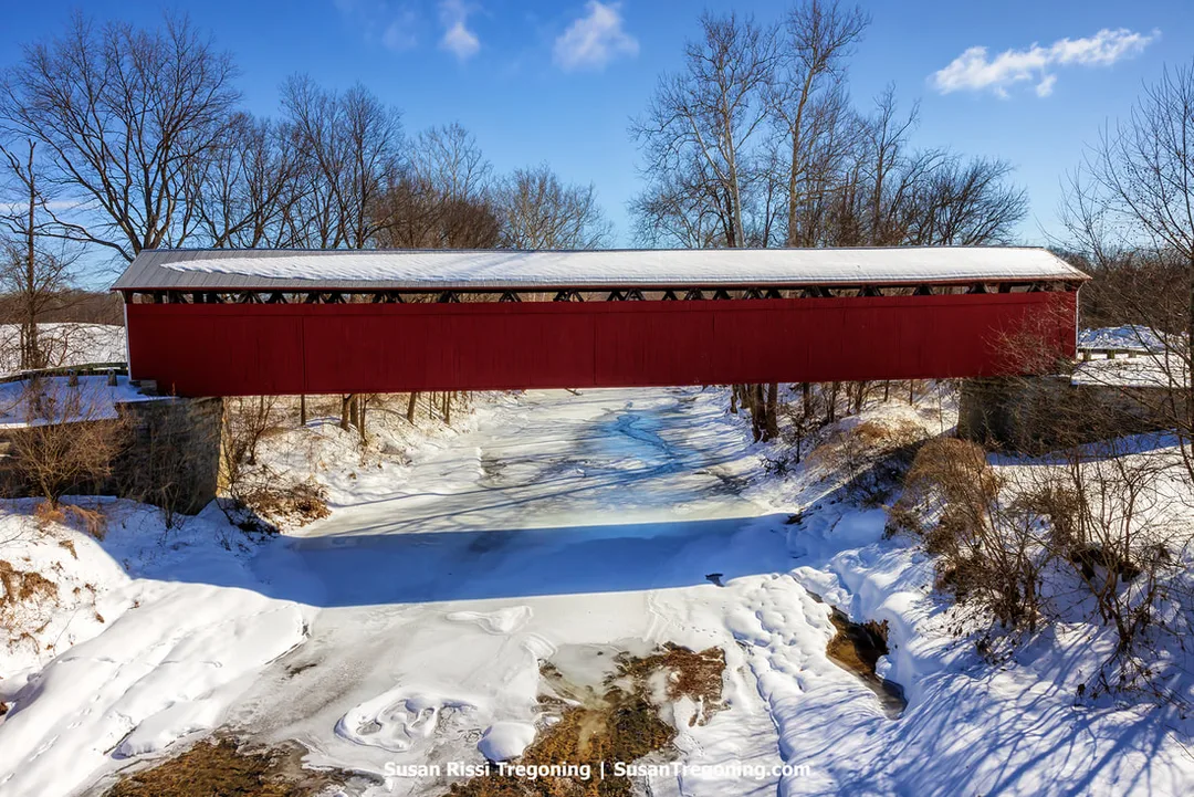 The historic red Scipio Covered Bridge in Jennings County, Indiana, shown broadside across the frozen Sand Creek, set in a bright, snow-covered winter landscape with leafless trees and clear sunlight.