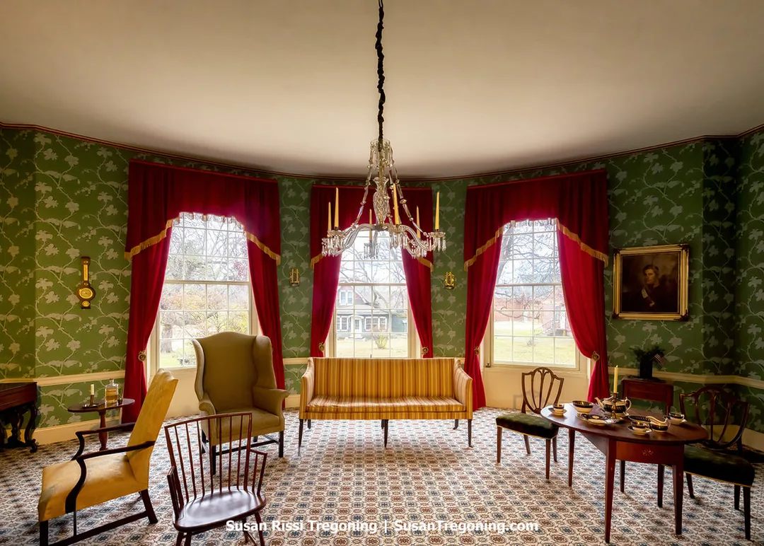 The formal parlor at Grouseland features green patterned wallpaper, red draperies with gold trim, and a crystal chandelier hanging above period furniture. A striped settee, upholstered chairs, and a wooden table set with fine china are arranged on a patterned carpet. A portrait in a gold frame hangs on the wall, and a wall clock is visible to the left. Large windows bring natural light into the historic room.