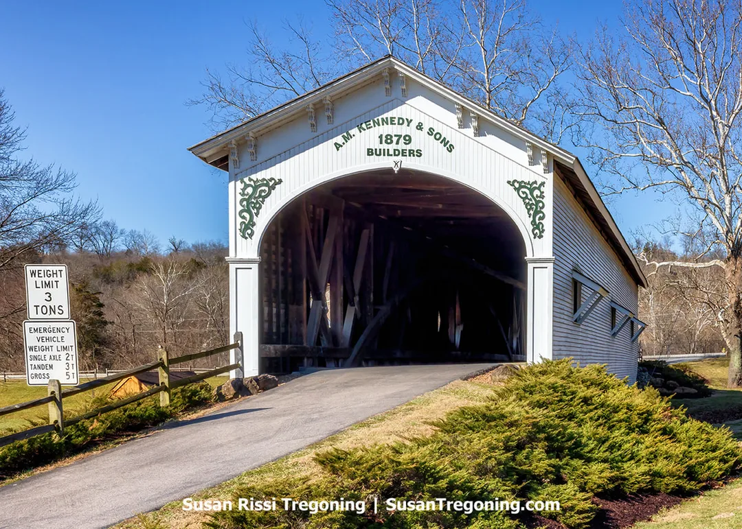 The historic Guilford white wooden covered bridge with green scrollwork above the entrance displays the inscription “A.M. Kennedy & Sons, 1879, Builders” in Guilford, Indiana. Leafless trees and shrubs surround the bridge, and a roadside sign to the left lists vehicle weight limits.