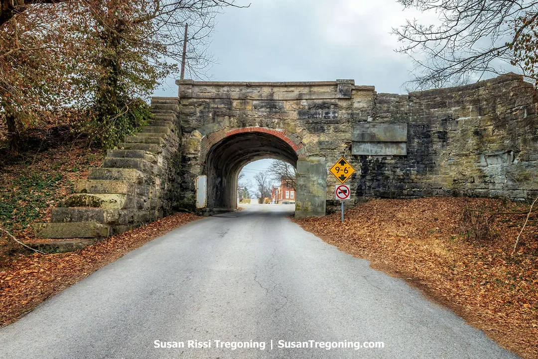 Historic limestone arch railroad bridge in Vernon, Indiana, photographed from the road that passes through it, showing the curved masonry arch framed by surrounding trees.