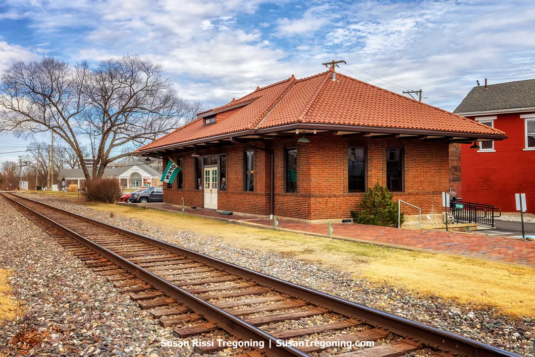 Railroad tracks still run alongside this historic red-brick train depot with a red-tiled roof in Aurora, Indiana, which now serves as the Aurora Public Library District's Local History Library. Leafless trees, parked cars, and nearby houses surround the building under a partly cloudy sky.