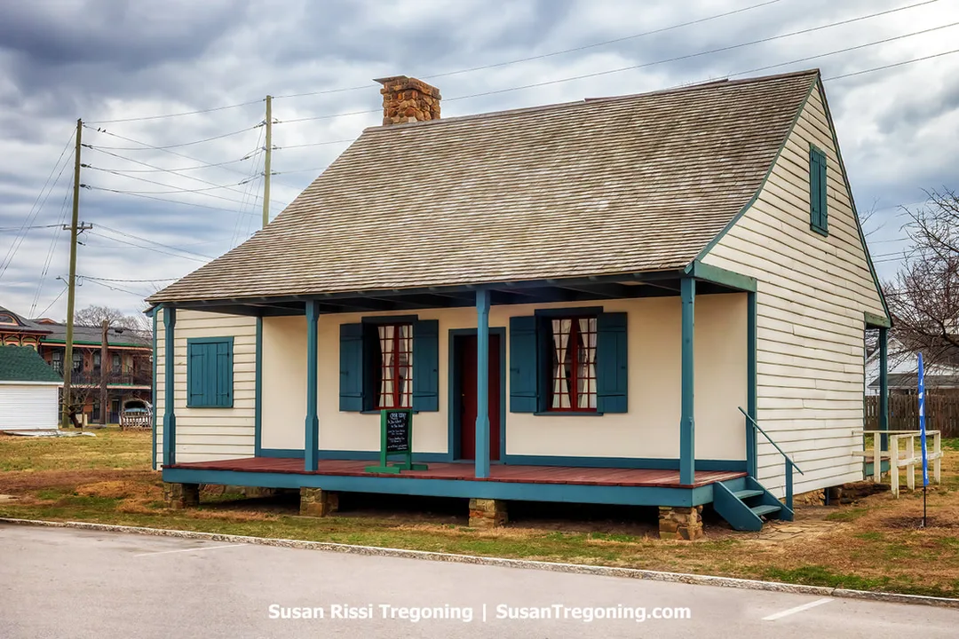 The Old French House, an authentic French Creole residence and the last surviving post‑on‑sill dwelling in Indiana, stands on a raised foundation with a steep roof, blue shutters, and a columned front porch. Nearby buildings and power lines are visible in the background.