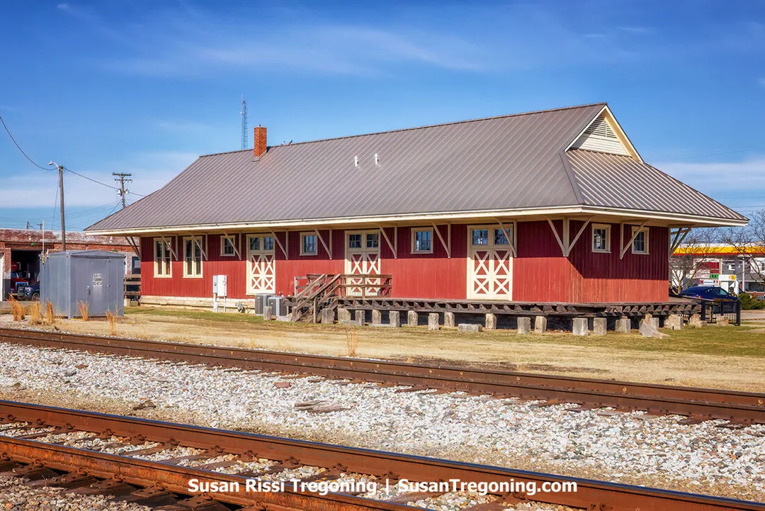 The Southern Indiana Railroad Freighthouse stands beside a set of train tracks on a clear day. The long, low wooden depot has a gabled roof, wide overhangs, and board‑and‑batten siding. A small platform runs along the track side of the building, and leafless trees rise behind it. The scene highlights the structure as the only surviving railroad depot in Seymour.