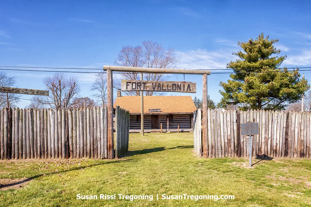 The entrance to the replica Fort Vallonia features a tall wooden palisade fence with a timber sign reading “Fort Vallonia” suspended between two posts. Inside the gate, a log cabin with a tan shingled roof is visible, marked as the garrison headquarters. Leafless trees, an evergreen, and a grassy lawn surround the fort under a bright blue sky.