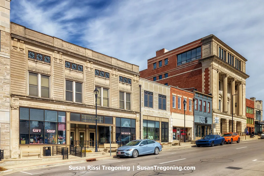 The Hamer‑Smith Building in downtown Bedford, Indiana, appears as a historic commercial structure with an ornate stone façade, large display windows, and signage for the Lawrence County Museum of History and its gift shop. The building sits along a street lined with other historic limestone storefronts, with a taller classical building visible in the background. Several parked cars line the curb beneath a partly cloudy sky.