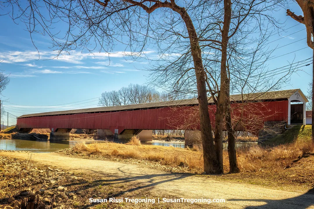A side-on view of the historic 1875 Medora Covered Bridge in Jackson County, Indiana. This long red wooden structure with a gray shingled roof spans the East Fork of the White River.