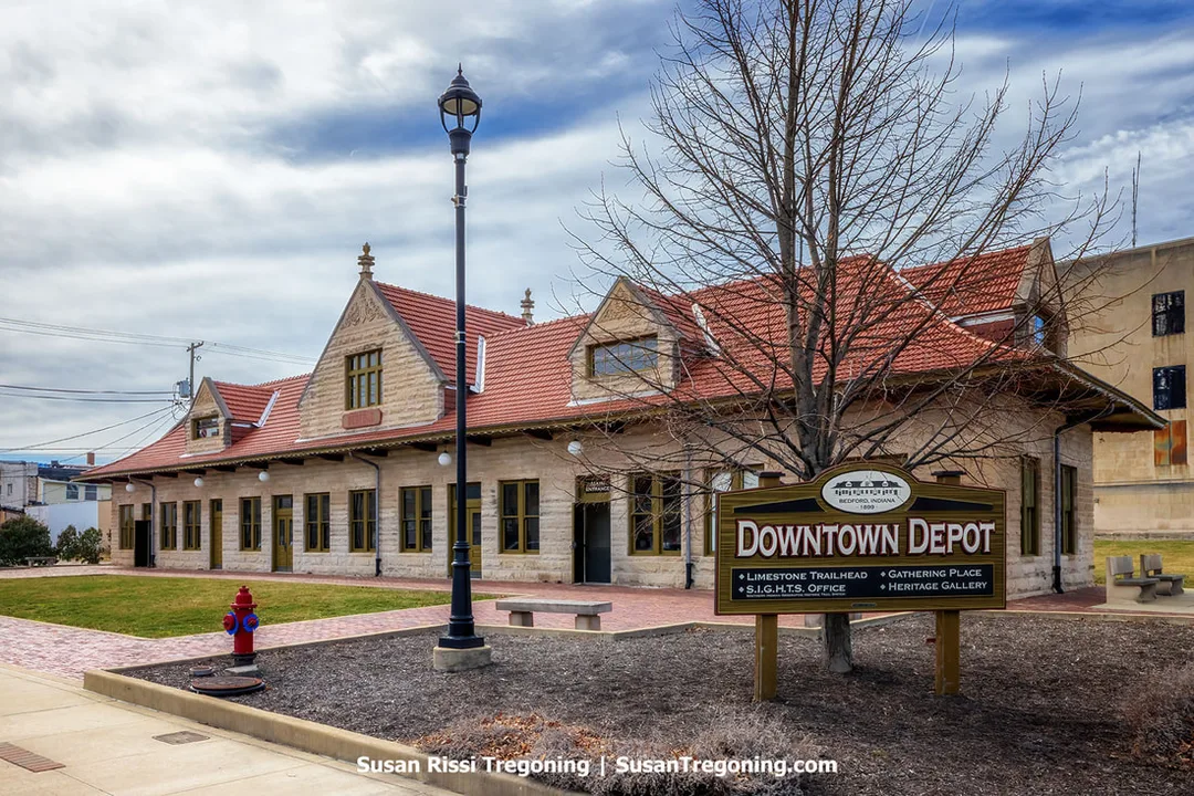 The restored Milwaukee Railroad Depot in Bedford, Indiana, is a single‑story limestone building with a red clay tile roof and gabled sections, shown on a partly cloudy day. A sign in front identifies it as the Downtown Depot and trailhead, with a leafless tree, streetlamp, fire hydrant, and bench in the foreground. Sidewalks and nearby buildings frame the historic structure.