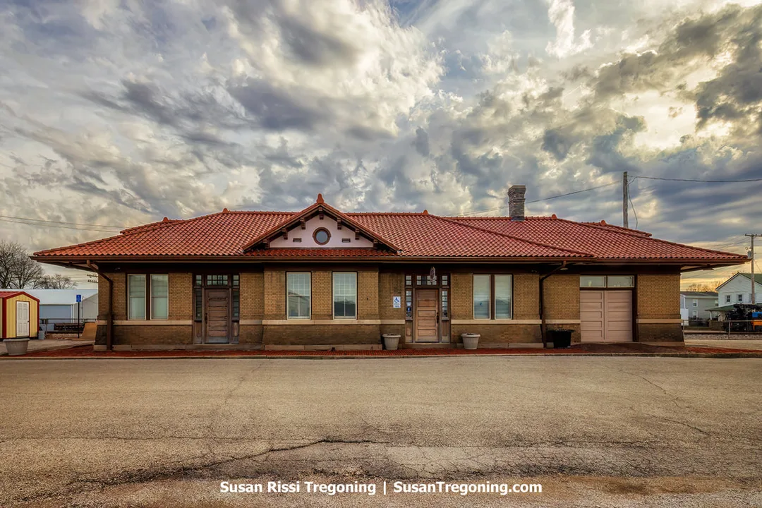 The restored 1906 B&O Southwestern Railroad Depot in Washington, Indiana, is a brick Mission Revival–style building with a red tile roof, symmetrical façade, and multiple windows beneath a dramatic, cloud‑filled sky. Two entrances sit along the front, one marked with an accessibility sign, and flower pots flank the doorways on the paved area in front.