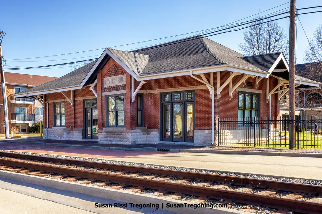 A historic Queen Anne-style red brick train station with a gabled roof and white decorative brackets stands beside a set of train tracks. A sign above the central entrance reads “Lynchburg.” Large windows and doors line the building, and a concrete platform runs along the tracks. A black metal fence borders the area, with nearby buildings and leafless trees visible under a clear blue winter sky.