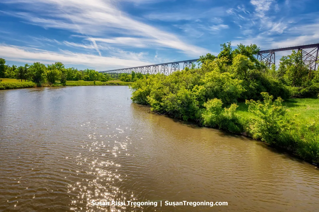 The Hi-Line Bridge spans the Sheyenne River near Valley City, North Dakota, with sunlight reflecting on the water and green trees lining the riverbank beneath a blue sky.