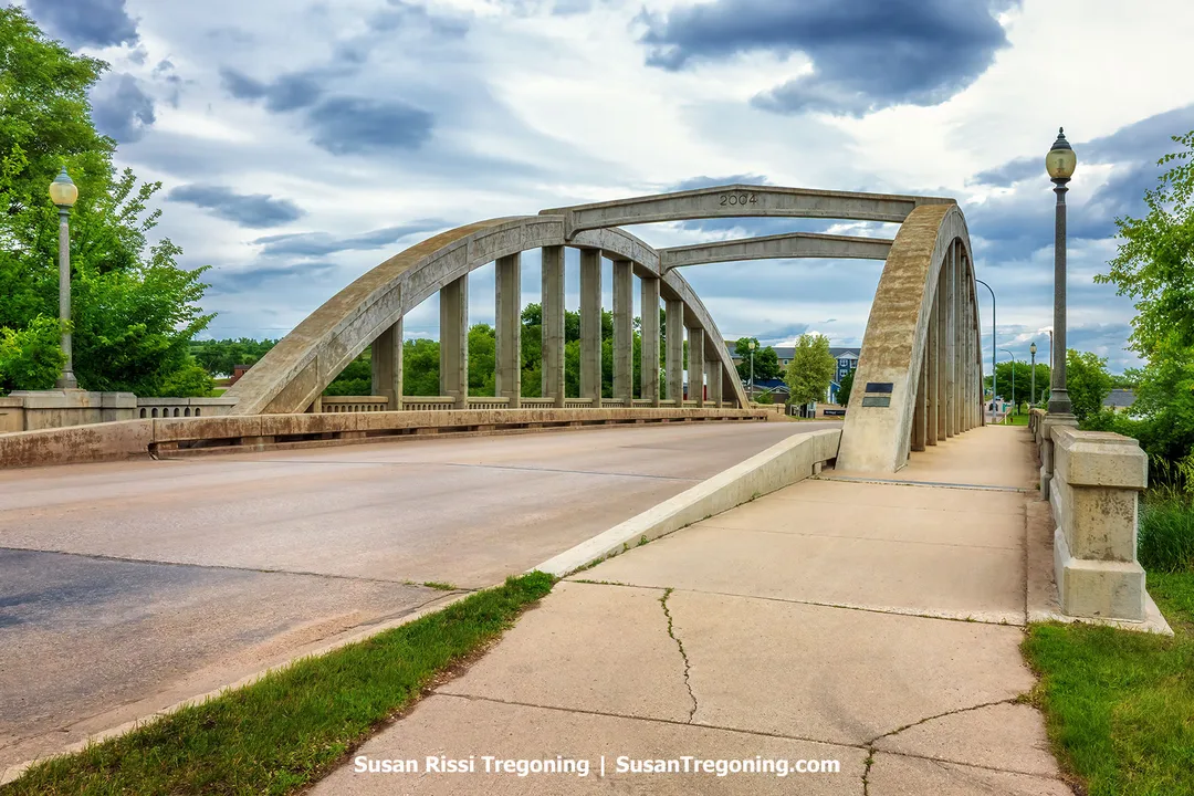 Rainbow (Main Street) Bridge over the Sheyenne River in Valley City, North Dakota, featuring graceful concrete through‑arches and lampposts under a dramatic sky. The bridge, rebuilt in 2004, preserves the historic Marsh Rainbow Arch design that made the original 1925‑26 structure unique in the state.