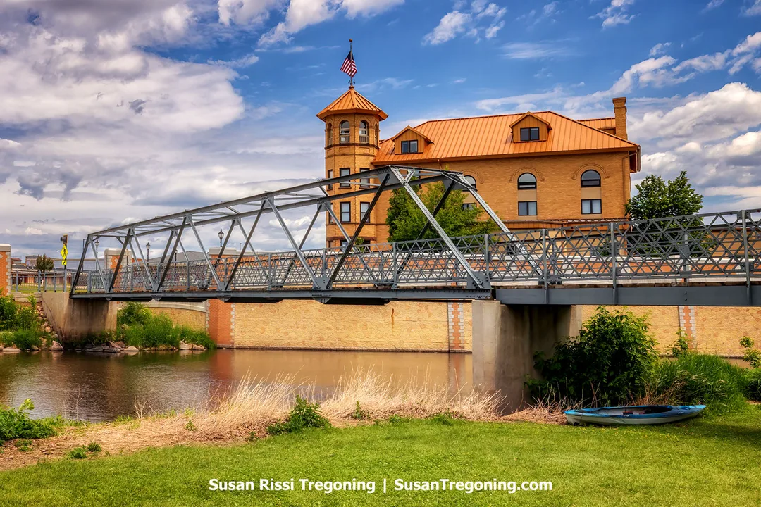 Historic City Park Footbridge spanning the Sheyenne River in Valley City, North Dakota, with the Elks Building’s orange-copper roof and turret rising behind it under a partly cloudy sky.