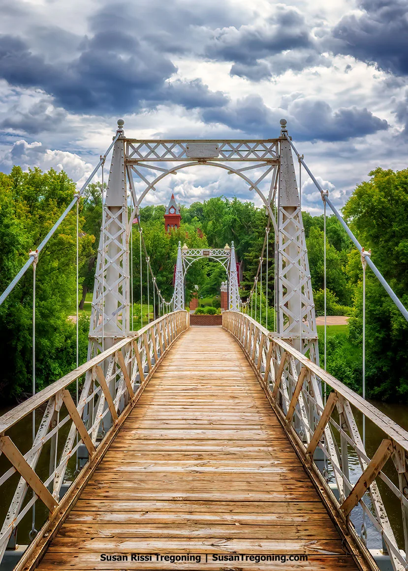 View across the Valley City State University Footbridge in Valley City, North Dakota, showing its wooden walkway and ornate steel towers under a dramatic sky, with the red‑brick clock tower of McFarland Hall rising beyond the trees.