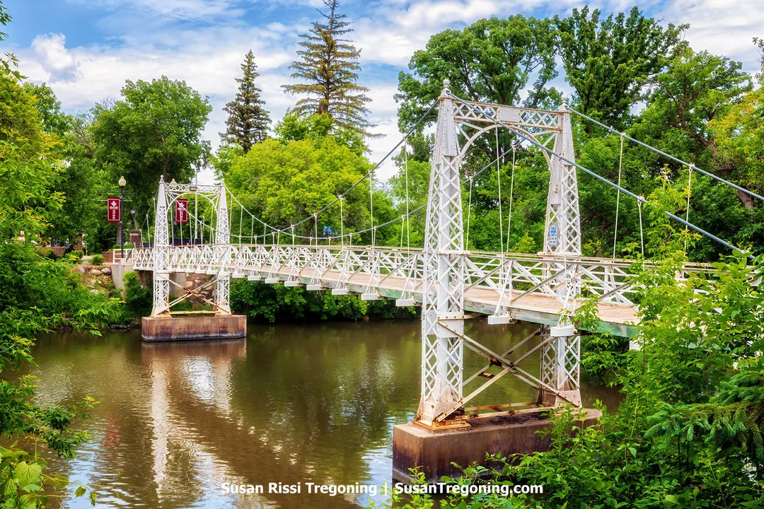 The Valley City State University Footbridge in Valley City, North Dakota — a white steel‑wire suspension bridge spanning the Sheyenne River, surrounded by lush summer greenery and reflected in calm water.