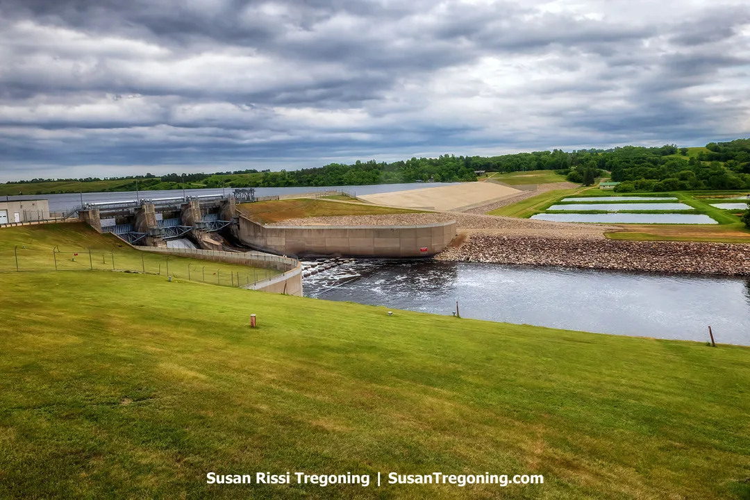 View of Baldhill Dam on the Sheyenne River in Barnes County, North Dakota. The earthen‑and‑concrete structure features three tainter gates releasing water downstream, with Lake Ashtabula visible behind the dam under a cloudy sky and green grassy slopes in the foreground.