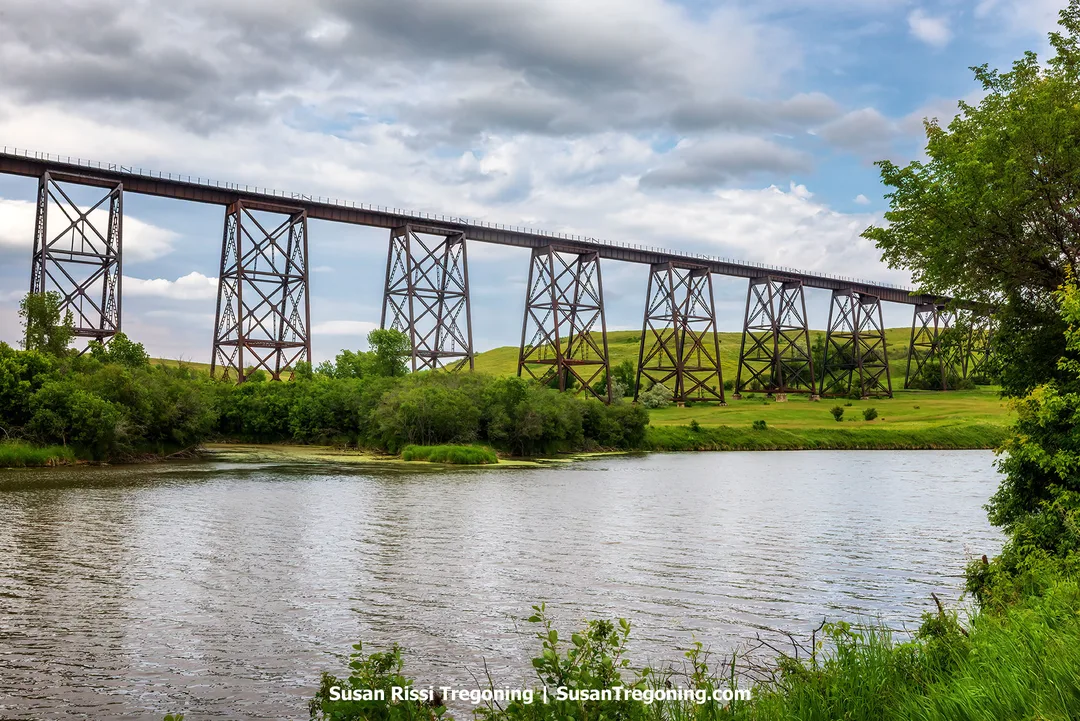 Hi Line Bridge, also known as the Valley City Viaduct, towering above the Sheyenne River Valley in Valley City, North Dakota. The 1908 steel railroad trestle stretches across green hills and trees, with its tall lattice framework reflected in the calm river below.