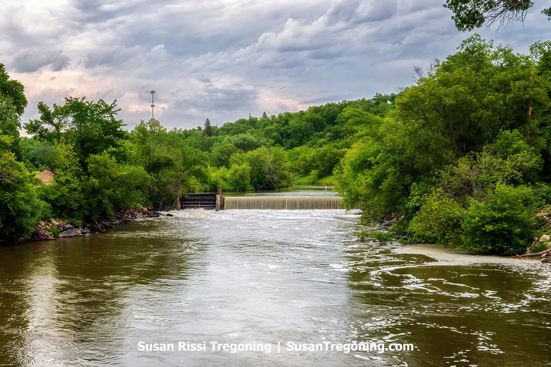 View of the Mill Bridge crossing the Sheyenne River in Valley City, North Dakota, near the old mill dam. The river flows calmly between lush green trees under a cloudy summer sky.