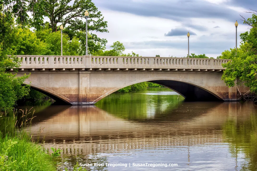 West City Park Bridge over the Sheyenne River in Valley City, North Dakota, featuring graceful concrete arches, decorative railings, and lampposts surrounded by lush green trees reflected in calm water.