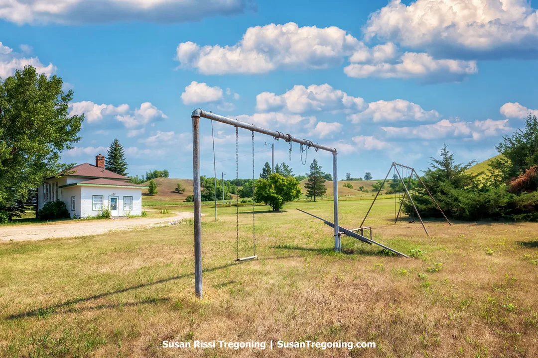 Historic King School stands beside an old playground with swings and a teeter-totter, surrounded by rolling prairie hills and open grassland in the Sheyenne River Valley of North Dakota.