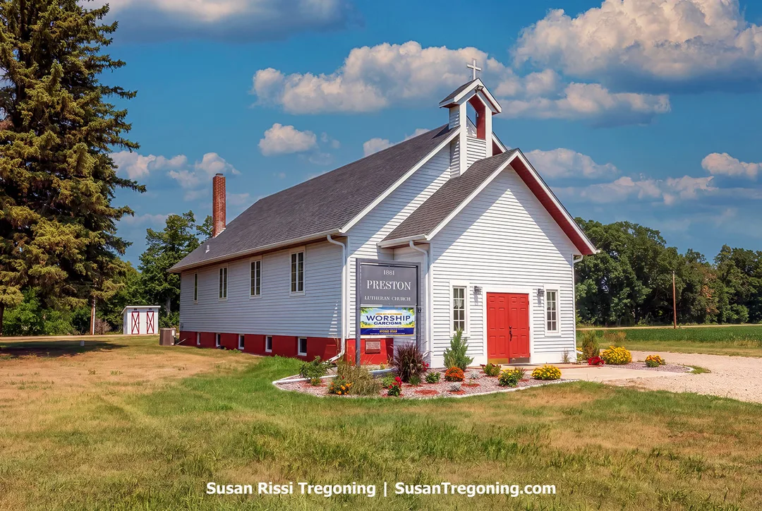 White wooden Preston Church with red doors and a bell tower, surrounded by grass and flowers along the Sheyenne River Valley National Scenic Byway in North Dakota.