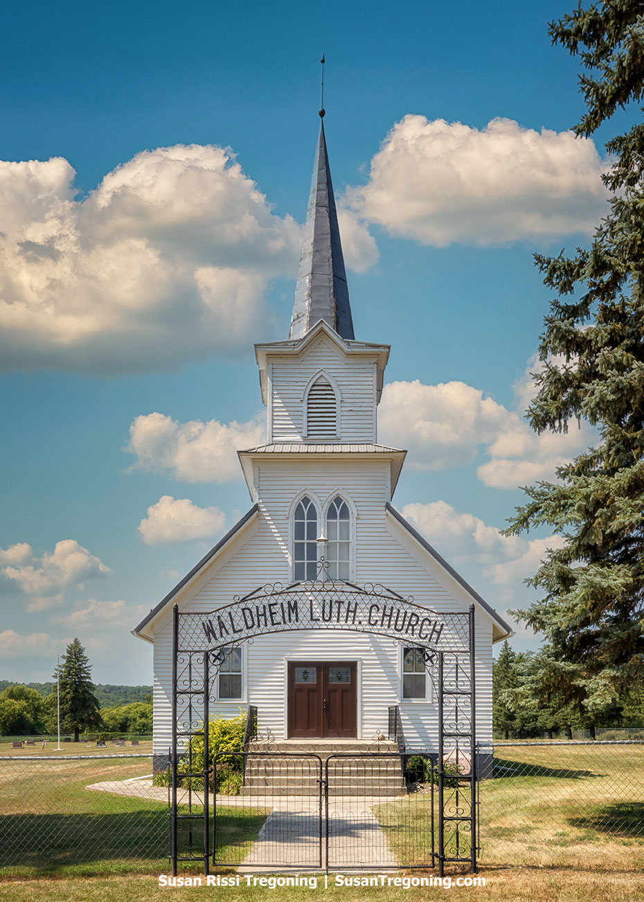 Front view of Waldheim Lutheran Church near Kathryn, North Dakota, featuring a white wood-frame exterior, tall steeple, and black iron entrance arch beneath a blue sky with large white clouds.