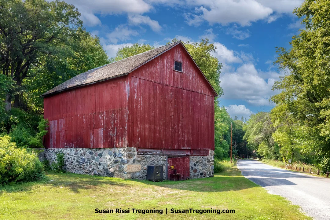Part of the T. J. Walker Historic District, this red barn with weathered wood siding and a stone foundation beside a narrow paved road bordered by trees near Fort Ransom, North Dakota.