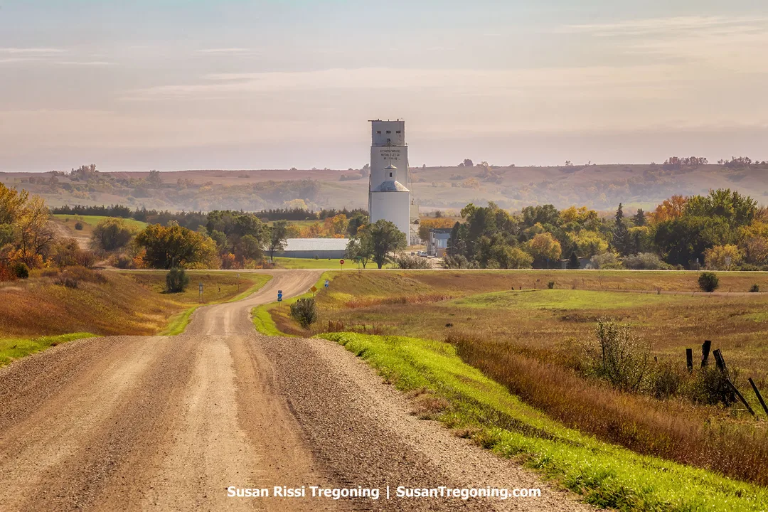 Rolling hills, prairie grass, and a gravel road leading toward Kathryn, North Dakota, with a grain elevator standing on the horizon beneath a soft autumn sky.