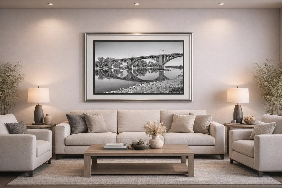 A warmly lit living room with a large beige sofa centered beneath a framed black‑and‑white photograph of the Lincoln Memorial Bridge in Vincennes, Indiana, reflected in calm water. Matching beige armchairs sit on either side of the sofa, with wooden end tables and identical table lamps beside them. A wooden coffee table holds a vase of dried flowers and decorative objects. Light-colored walls, recessed ceiling lights, and tall potted plants in the corners create a balanced, serene space.