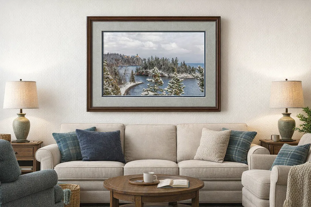Framed winter landscape wall art featuring a snow‑covered shoreline, evergreen trees, and a lighthouse overlooking calm blue water. Displayed above a beige sofa in a cozy living room with warm lighting, wood accents, and blue‑green pillows. The piece is presented in a dark wood frame with layered blue‑gray mats, creating a rustic, inviting atmosphere that complements the art’s serene northern feel.