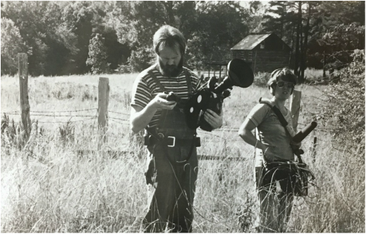In a field, Peter Adair looks at light meter, holding a film camera, Lucy Phenix holds a recorder and mic.