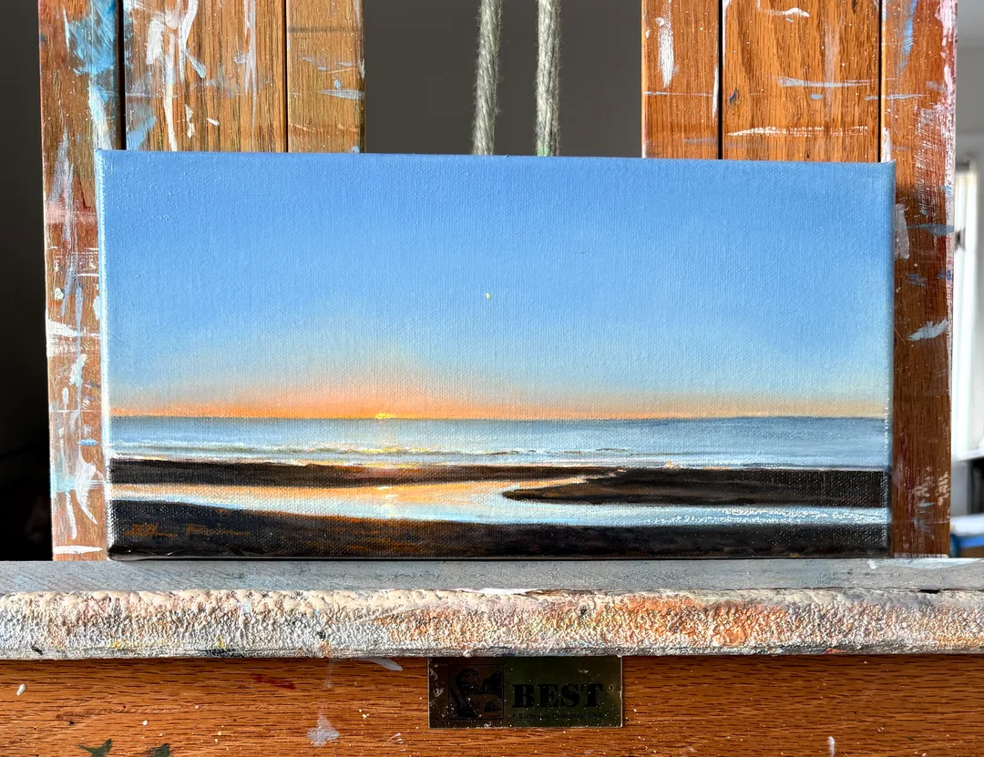 A radiant, small, oil painting of the sun rising over the Atlantic ocean, the sky gradually going from blazing on the horizon to deep blues, all reflected in a tidal pool in the foreground sand.