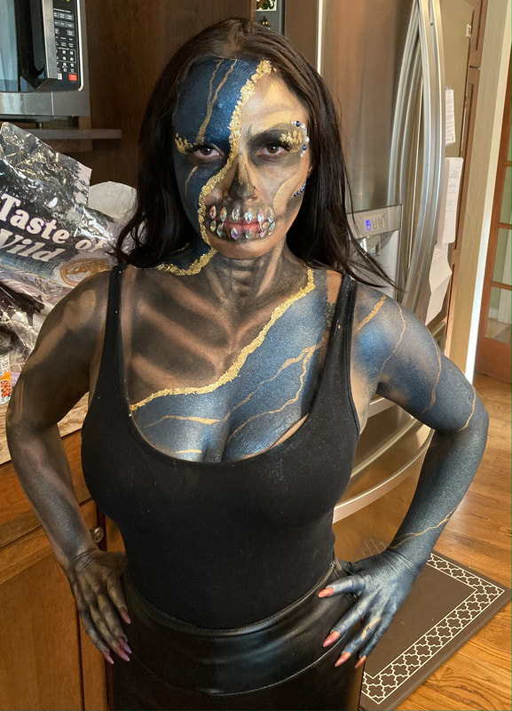 A young woman wears blue and gold skull face and body painting with diamond teeth while standing in a kitchen, San Francisco