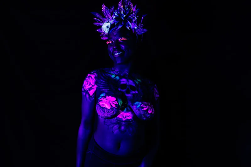 A young black woman in San Francisco, Ca, model's a UV reactive body painting and blacklight reactive eyelashes. The design splashes across her chest, a skull in the center with wing's spreading out following her collar bone. Glowing pink roses and white mandala's surround the design. She also sports a spikey gold and black floral crown.