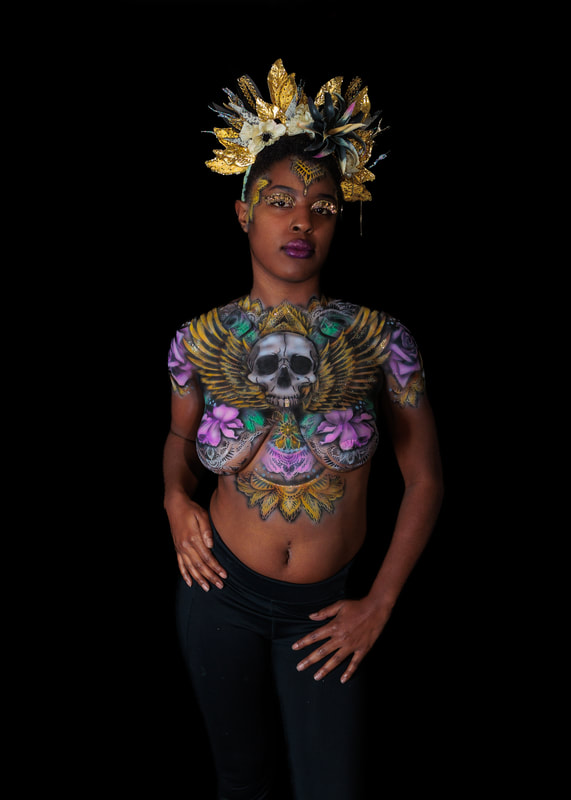 A young black woman in San Francisco, Ca, model's a UV reactive body painting and blacklight reactive eyelashes. The design splashes across her chest, a skull in the center with wing's spreading out following her collar bone. Glowing pink roses and white mandala's surround the design. She also sports a spikey gold and black floral crown.