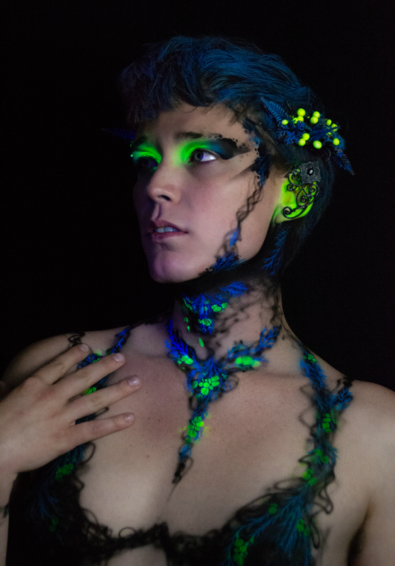 A young white woman in Oakland, Ca is wearing UV reactive body paint and blacklight activated makeup. She resembles a fairy with a head piece and pointed ear cuff's, but the design in inspired by fungus and rot. Black tendrils sweep up around her neck like a necklace and top. Green berries and blue plant matter glow in the dark.
