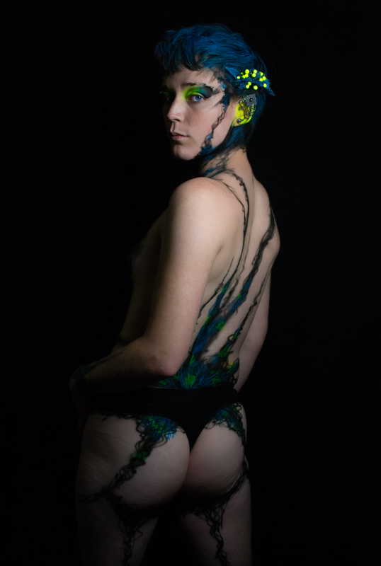 A young white woman in Oakland, Ca is wearing UV reactive body paint and blacklight activated makeup. She resembles a fairy with a head piece and pointed ear cuff's, but the design in inspired by fungus and rot. Black tendrils sweep up around her neck like a necklace and top. Green berries and blue plant matter glow in the dark.