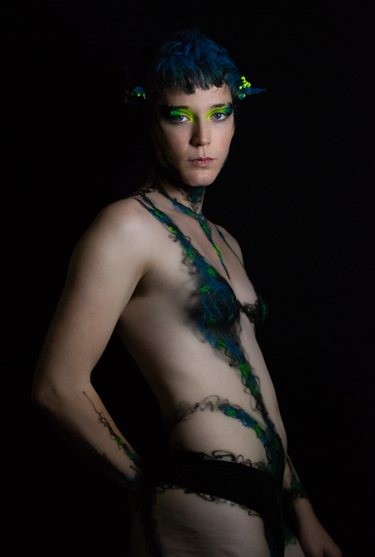 A young white woman in Oakland, Ca is wearing UV reactive body paint and blacklight activated makeup. She resembles a fairy with a head piece and pointed ear cuff's, but the design in inspired by fungus and rot. Black tendrils sweep up around her neck like a necklace and top. Green berries and blue plant matter glow in the dark.