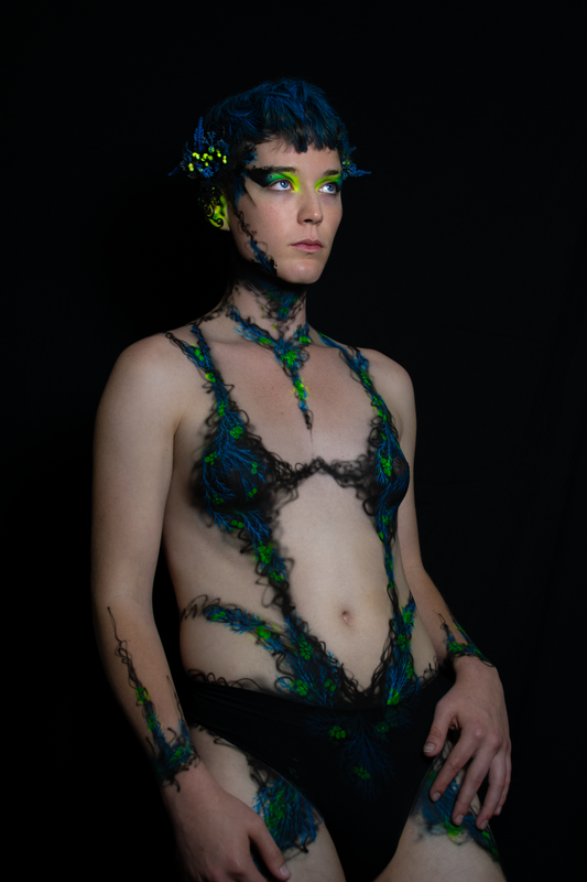 A young white woman in Oakland, Ca is wearing UV reactive body paint and blacklight activated makeup. She resembles a fairy with a head piece and pointed ear cuff's, but the design in inspired by fungus and rot. Black tendrils sweep up around her neck like a necklace and top. Green berries and blue plant matter glow in the dark.