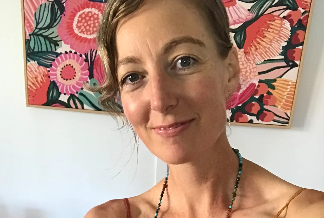 A smiling person with dark blonde hair and kind eyes.  Their hair is pulled back and fringe sweeps to one side in light waves.  They are wearing a colourful beaded necklace and stand in front of a colourful abstract picture of native Australian flowers in pinks, oranges and greens.