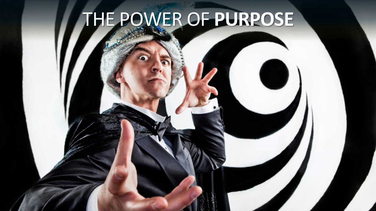 A performer costumed as a stage hypnotist, in a sparkly silver turban with a feather, black tuxedo, bow tie and velvet cape, stares wide-eyed at the camera. One hand is raised beside his head and the other reaches towards the viewer, in front of a black-and-white hypnotic spiral. Headline reads “The power of purpose”, with “purpose” in bold.