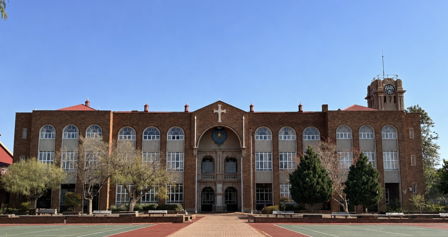Front view of St Patrick’s CBC Kimberley, showing the brick College building with its central entrance, arched windows and distinctive War Memorial Clock tower rising above the right-hand side. Tennis courts are visible in the foreground.