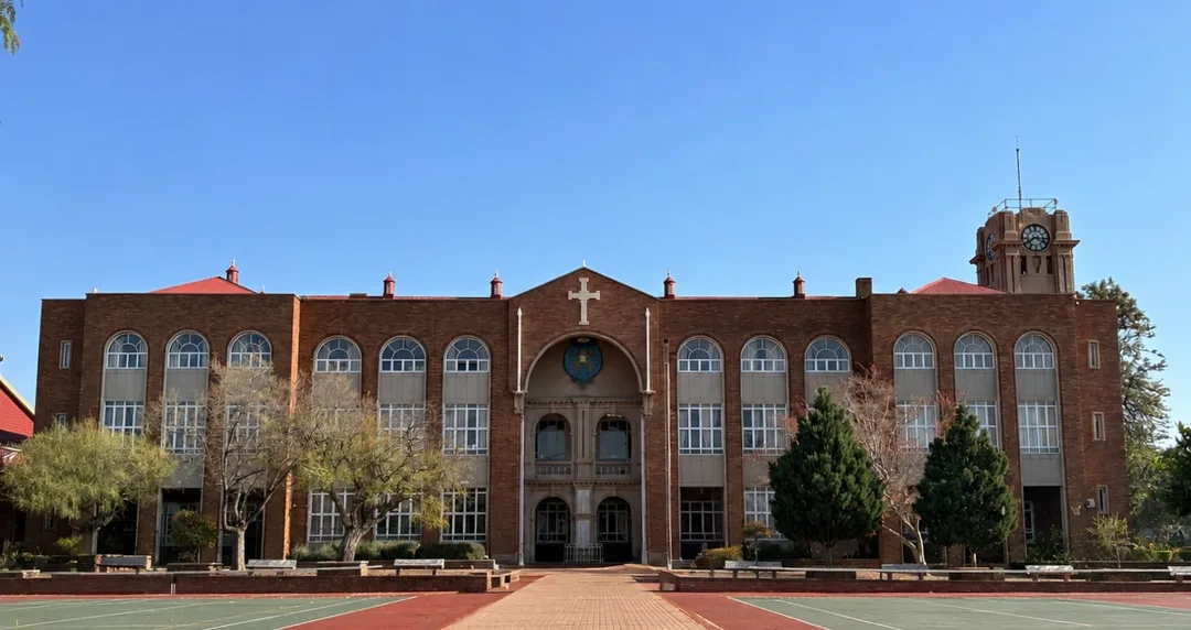Front view of St Patrick’s CBC Kimberley, showing the brick College building with its central entrance, arched windows and distinctive War Memorial Clock tower rising above the right-hand side. Tennis courts are visible in the foreground.